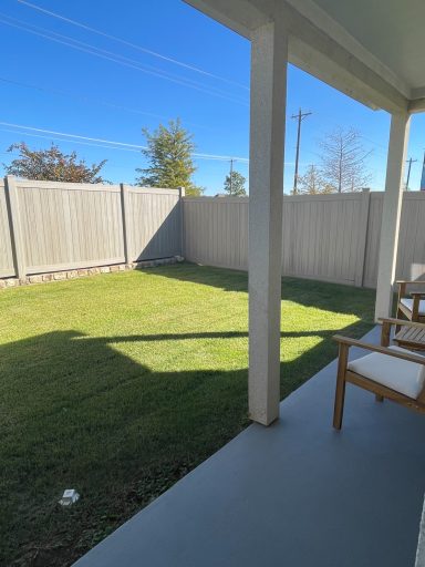 View of a grassy backyard enclosed by a fence, with a patio and two chairs.